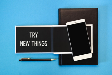 A white notebook with black pages, a smartphone and a pen on a blue background. The inscription TRY NEW THINGS