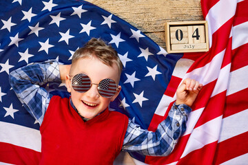 Cute and cheerful guy lying on a wooden background next to the American flag with glasses. Independence - obrazy, fototapety, plakaty