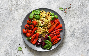 Grilled vegetables: zucchini, paprika, tomatoes, onions in a large plate on a concrete kitchen table.