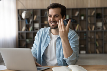 Smiling businessman recording audio voice message on smartphone, speaking, sitting at work desk with laptop, positive young man chatting online by speakerphone, activating digital assistant on gadget