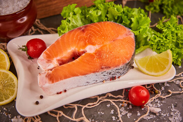 Raw salmon steak on a slate-black background with a fresh green salad, surrounded by lemon slices, cherry tomatoes and sea salt. The concept of healthy food and fresh seafood and delicacies.