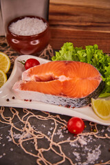 Raw salmon steak on a slate-black background with a fresh green salad, surrounded by lemon slices, cherry tomatoes and sea salt. The concept of healthy food and fresh seafood and delicacies.