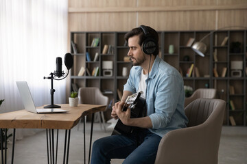 Confident man in headphones playing acoustic guitar, looking at laptop screen, sitting at desk with microphone, musician artist recording new song in home studio, student practicing, online course