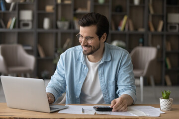 Smiling businessman in glasses using laptop, calculating bills, managing finances, happy young man planning budget, sitting at desk at home, browsing online banking service, satisfied by money refund - obrazy, fototapety, plakaty
