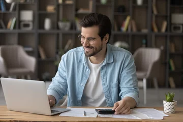 Fotobehang Smiling businessman in glasses using laptop, calculating bills, managing finances, happy young man planning budget, sitting at desk at home, browsing online banking service, satisfied by money refund © fizkes