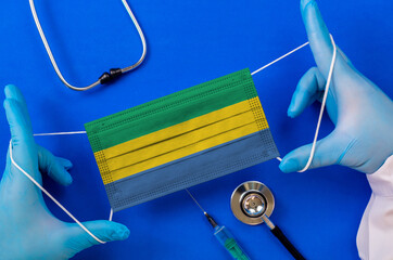 Medical mask with the flag of Gabon in the hands of a doctor and stethoscope, flat lay. Coronavirus...