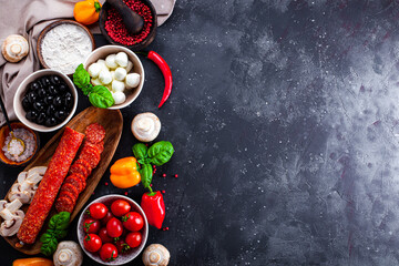  Pizza ingredients on the dark gray background, free space for text. Pepperoni sausage, mozzarella cheese, tomatoes, olives, mushrooms and flour are different products for making pizza and pasta