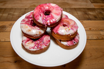 A selection of oil fried doughnuts topped with sweet pink and white icing and sprinkles on a white plate on a wooden kitchen table