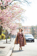 Fototapeta premium In the spring, a woman walks along a blooming street with sakura trees. A girl in a long silk elegant vintage dress walks among the flowering trees