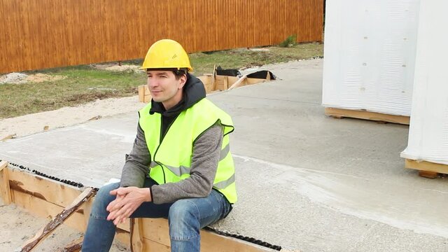 A construction worker in a protective yellow hardhat and a signal vest sit down in foundation with wooden formwork and aerated concrete blocks on a construction site. Foreman, building materials.