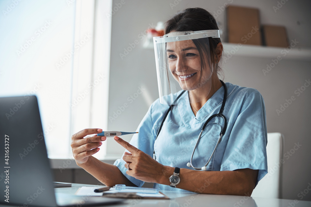 Wall mural Joyful doctor pointing at thermometer during online consultation