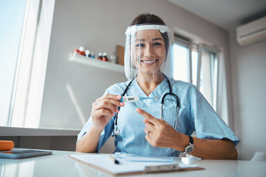 Cheerful doctor in face mask pointing at digital thermometer