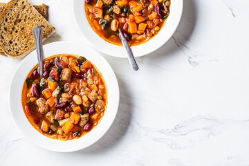 Beans soup with sausages in white plate, top view, white marble background. Italian cuisine concept.
