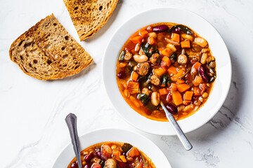 Beans soup with sausages in white plate, top view, white marble background. Italian cuisine concept.