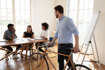 Young male manager came with bicycle, looking at mixed race diverse creative teammates discussing...
