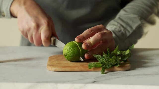 Bartender is cutting a juicy green lime on a chopping board with a knife, and a sprig of mint is lying next to it.