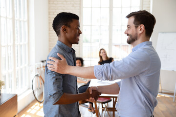 Smiling young male boss leader shaking hands with millennial african ethnicity intern employee,...
