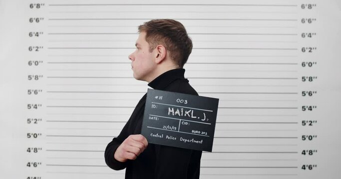Mugshot of millennial guy turning to sides while holding sign and posing for photo . Crop view of criminal man in black turtleneck looking to camera in police department. Concept of crime.