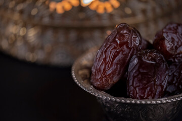 Religious Islamic festival and holy month of Ramadan, EID KAREEM, concept: Close up on a metallic bowl with dried date fruits and a golden lantern with candle light on dark background with copy space