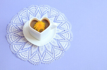 A cup of coffee with a dandelion flower in it on a white lace napkin on a blue background, side view, space for text