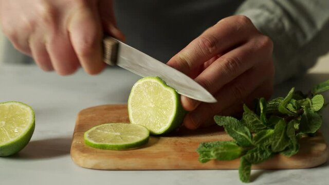 Bartender is cutting a juicy green lime on a chopping board with a knife, and a sprig of mint is lying next to it.