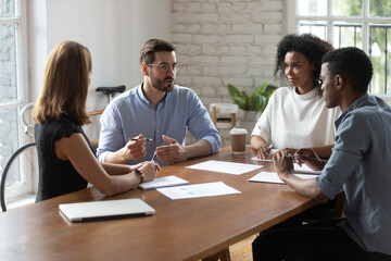 Concentrated young male team leader in eyeglasses instructing multiracial employees at briefing meeting, sitting at table in modern office. Skilled mixed race colleagues discussing project indoors.