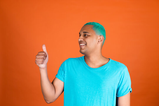 Black African Man In Casual On Orange Background Blue Hair Happy Positive Smile Looking Left To Empty Space  Shows Thumb Up