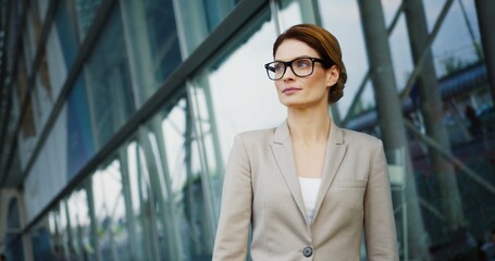 Portrait shot of the Caucasian pretty business lady smiling to the camera and crossing her hands while standing on the glass building background. Outside