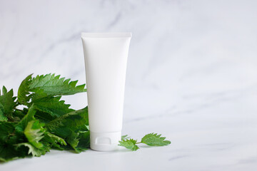 Obraz na płótnie Canvas Alternative nature cosmetics concept. White cream tube with nettle plant leaves on marble background. Mockup for packaging product