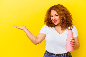 Young latin woman holding a thermos isolated on yellow background showing a copy space on a palm and holding another hand on waist.