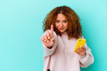Young latin woman holding mobile phone isolated on blue background showing number one with finger.
