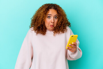 Young latin woman holding mobile phone isolated on blue background shrugs shoulders and open eyes...