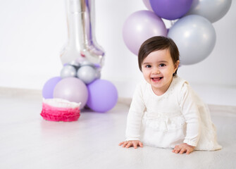 baby girl in white dress sitting on the floor celebrating her first birthday with cake and balloons.