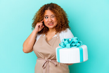 Young pastry latin woman holding a cake isolated on blue background covering ears with hands.