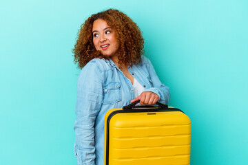 Young latin traveler curvy woman holding a suitcase isolated on blue background looks aside...