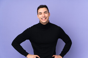 Young caucasian handsome man with turtleneck sweater isolated on purple background posing with arms at hip and smiling