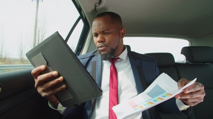 Successful handsome african american businessman in formalwear networking on digital tablet, analyzing financial data and charts while riding in backseat of car during business travel. - Powered by Adobe