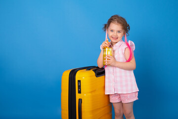 Obraz premium Funny child girl in pink clothes holding glass of orange juice with yellow suitcase on blue background
