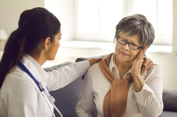 Obraz premium Mature woman telling doctor about pains and severe headaches during home visit or health checkup at hospital. Medical specialist helping older female patient who is suffering from stress and weakness