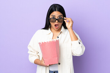 Young latin woman woman isolated on white background surprised with 3d glasses and holding a big bucket of popcorns