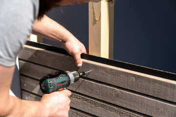 tough construction worker applies wooden siding to a garage and drills screws - obrazy, fototapety, plakaty