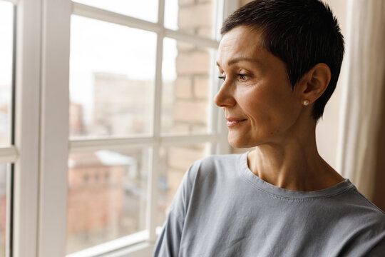 Close up image of beautiful short haired mature female with face wrinkles relaxing indoors, standing by window, daydreaming, having thoughtful look. Attractive retired woman spending time at home