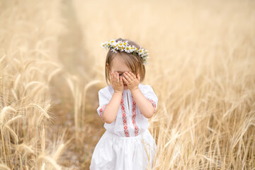 A little girl in a national Belarusian shirt covers her face with her hands. A child in a golden...