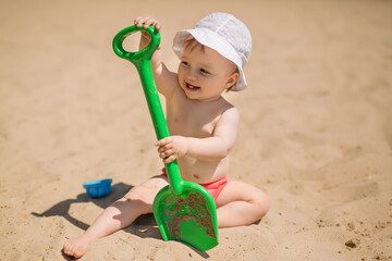 Baby with a big green toy shovel on the beach on a hot day. Sunscreen for children. A child in a panama hat and panties plays in the sand