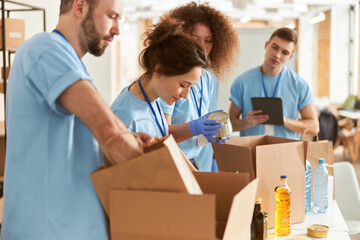 Diverse volunteers in protective gloves sorting, packing canned food in cardboard boxes, working...