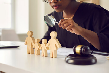 Black judge or lawyer looking in magnifying glass at little mom, dad children figures on desk....