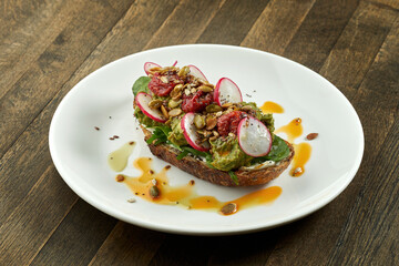Avocado toast with guacamole, radish, sun-dried tomatoes and seeds on rye bread, served in a white plate on a wooden background. Vegetarian food