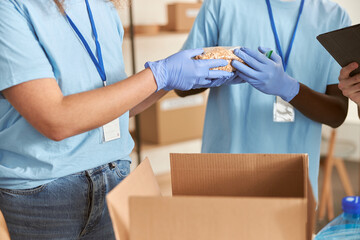 Close up shot of hands of volunteers in protective gloves sorting, packing food in cardboard box,...