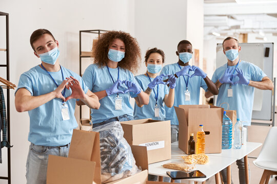 Group Of Diverse People Wearing Blue Uniform, Protective Masks And Gloves Showing Love Heart Sign While Volunteering In Community, Sorting Donated Food Items