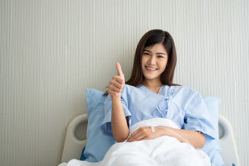 Happy Asian female patient smiling, lies on the bed, and raises thumb up to show confidence in...